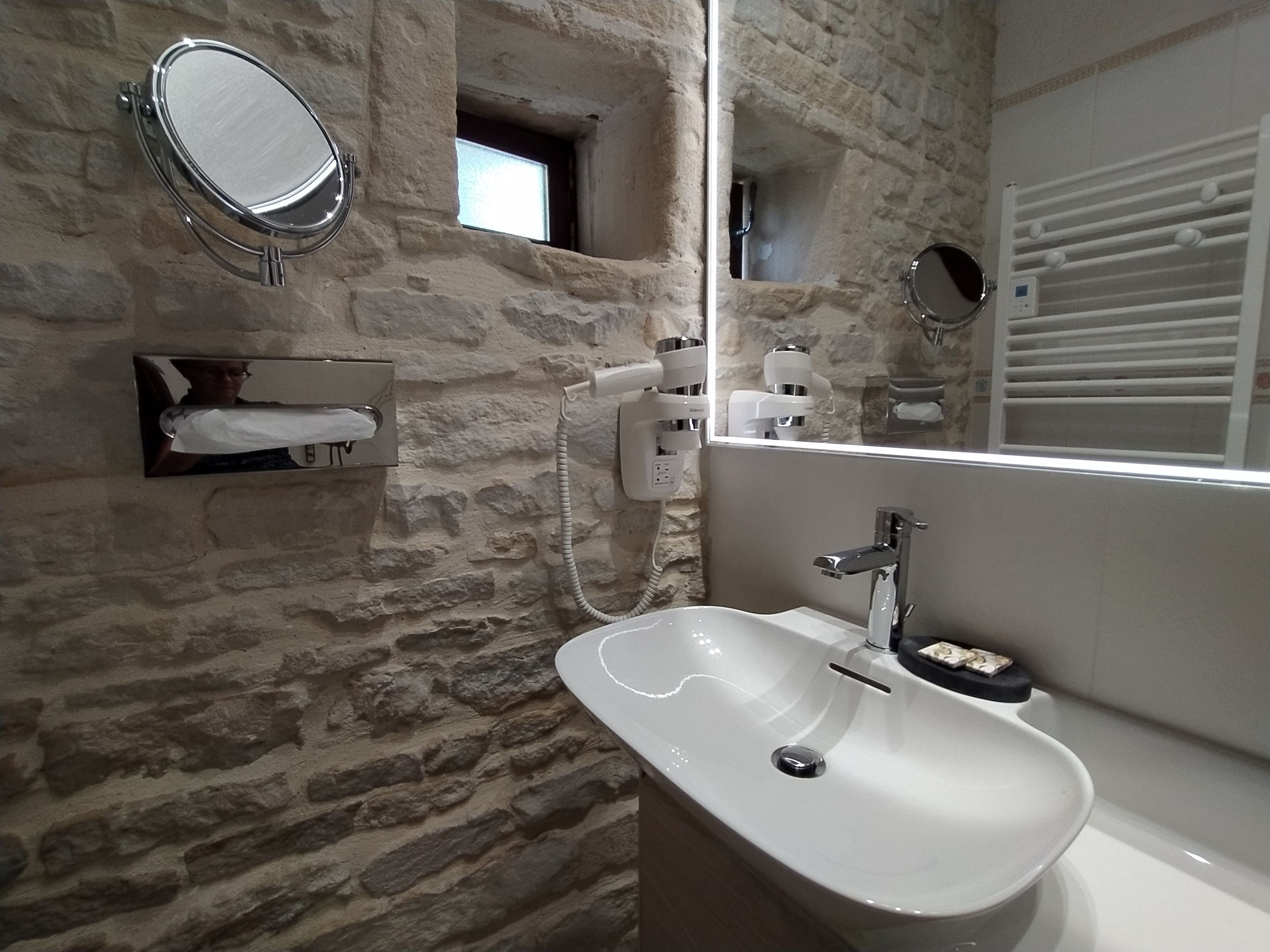 A bathroom with a stone wall, a white sink, a mirror, and a towel dispenser.