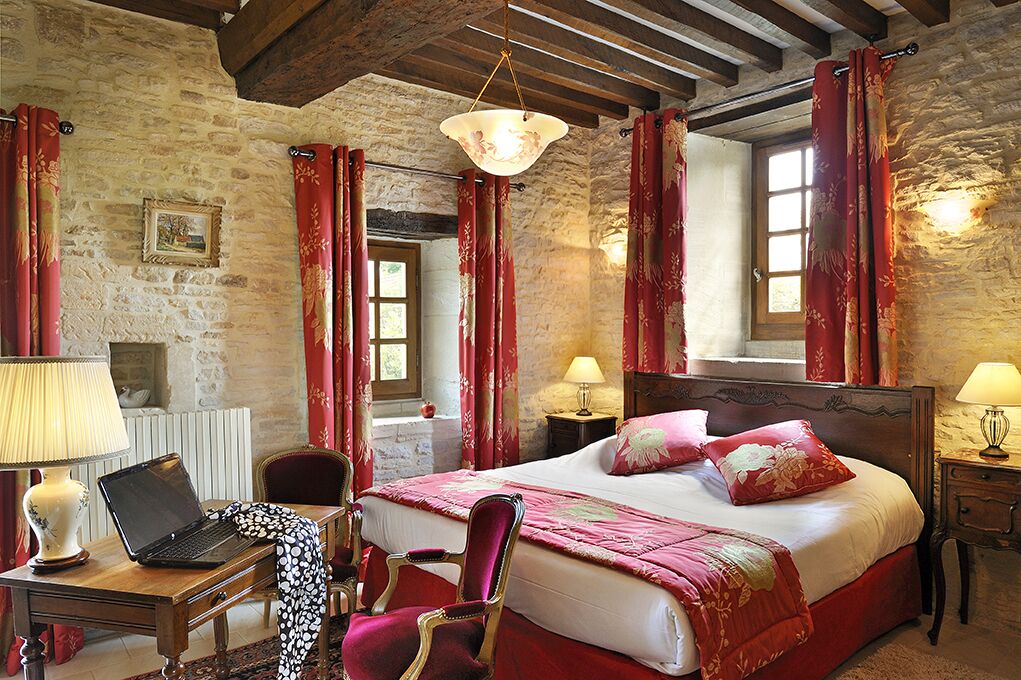 A bedroom with a stone wall, wooden ceiling beams, a bed with red bedding, a desk with a laptop, and two bedside lamps.