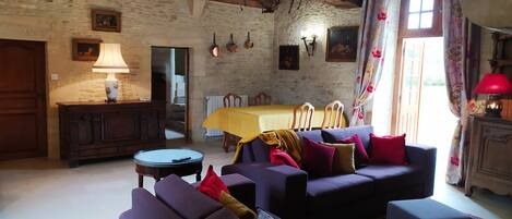 A rustic living room with a stone wall, wooden beams, a round coffee table, purple sofas, and a dining area with a yellow tablecloth.