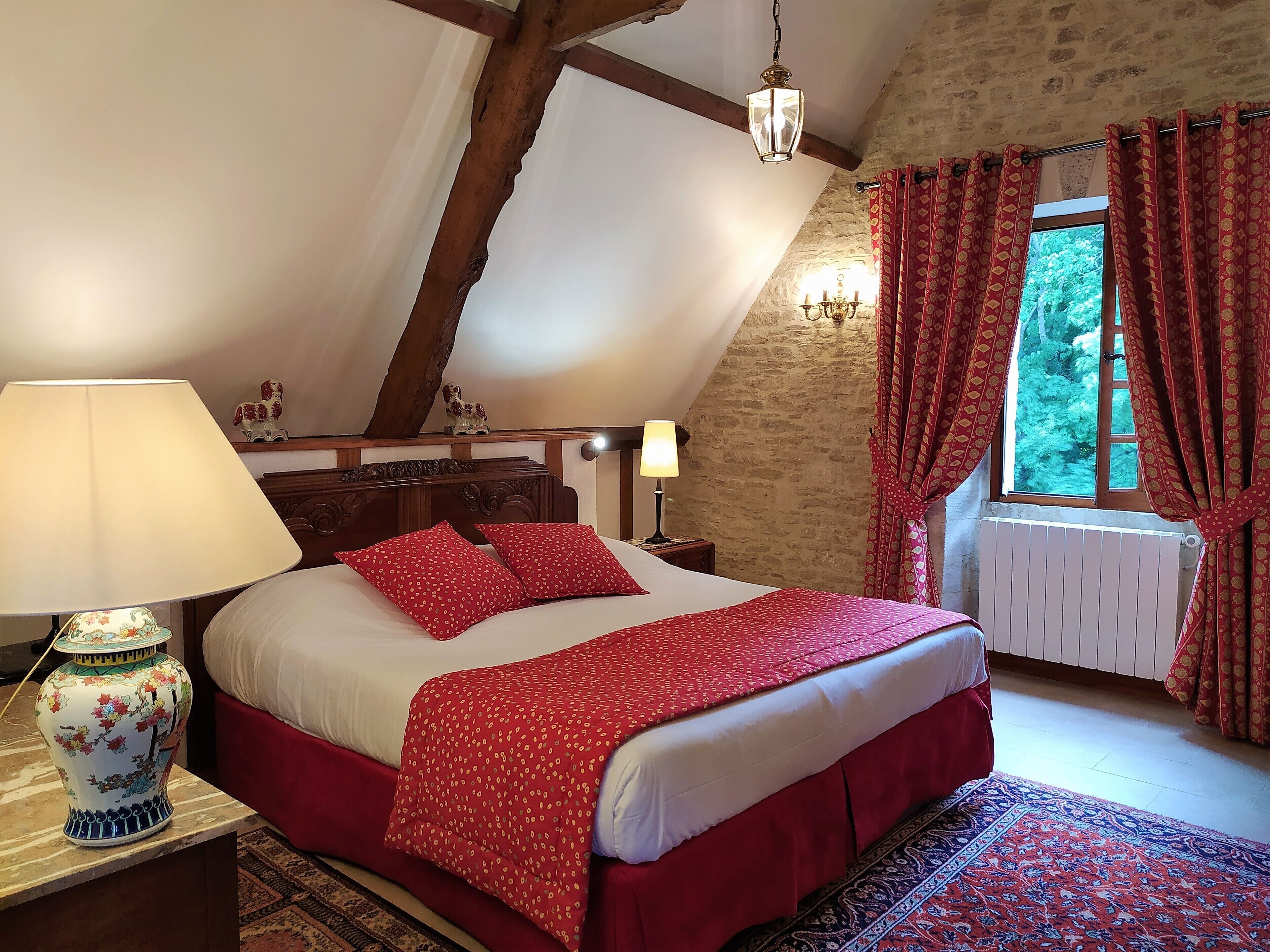 Un dormitorio con techo inclinado, una cama con ropa de cama roja, una lámpara decorativa y vistas a árboles a través de una ventana.