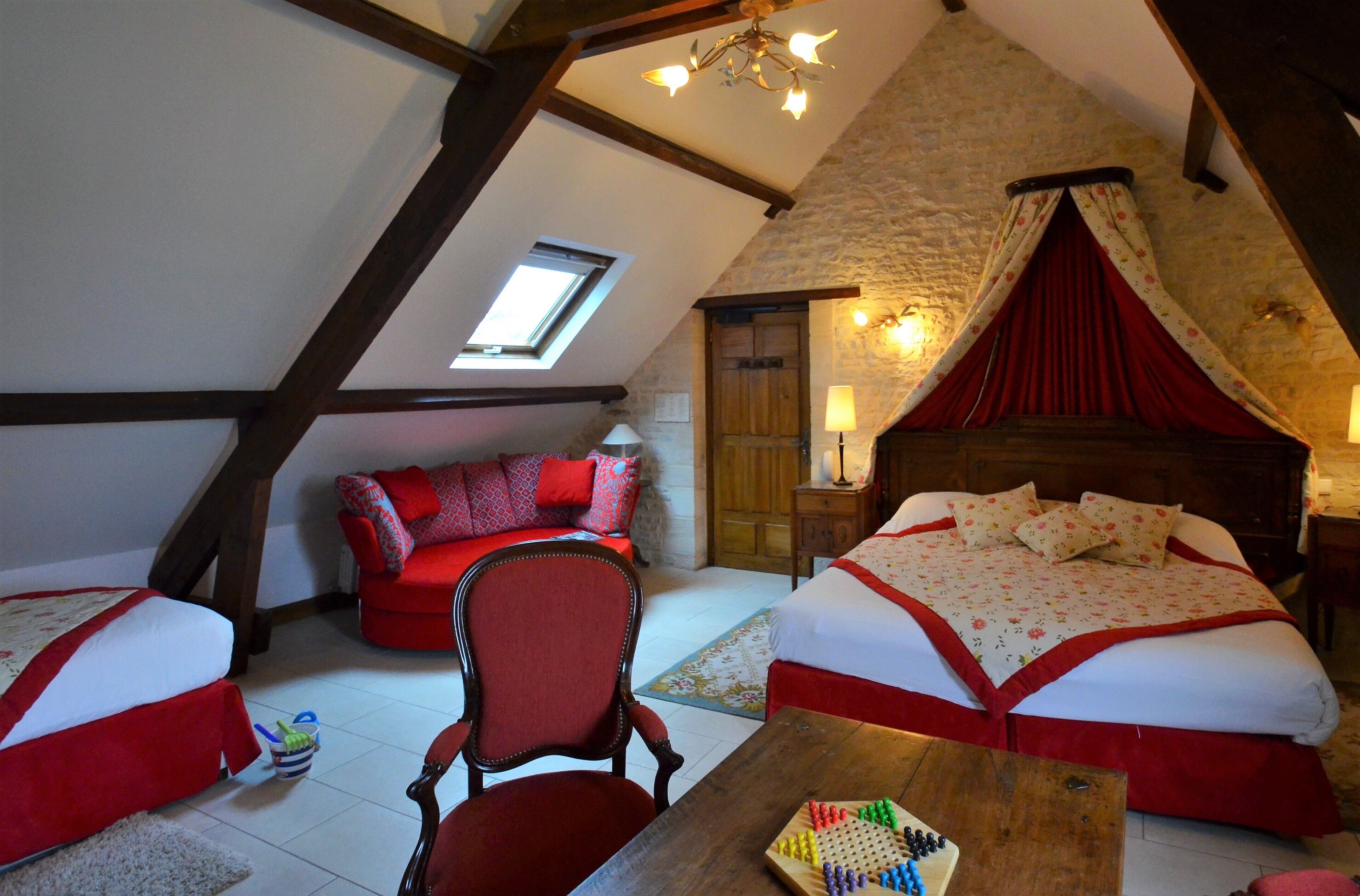 A cozy attic bedroom with a sloped ceiling, a wooden bed with red bedding, a red chair, and a small table with a board game.