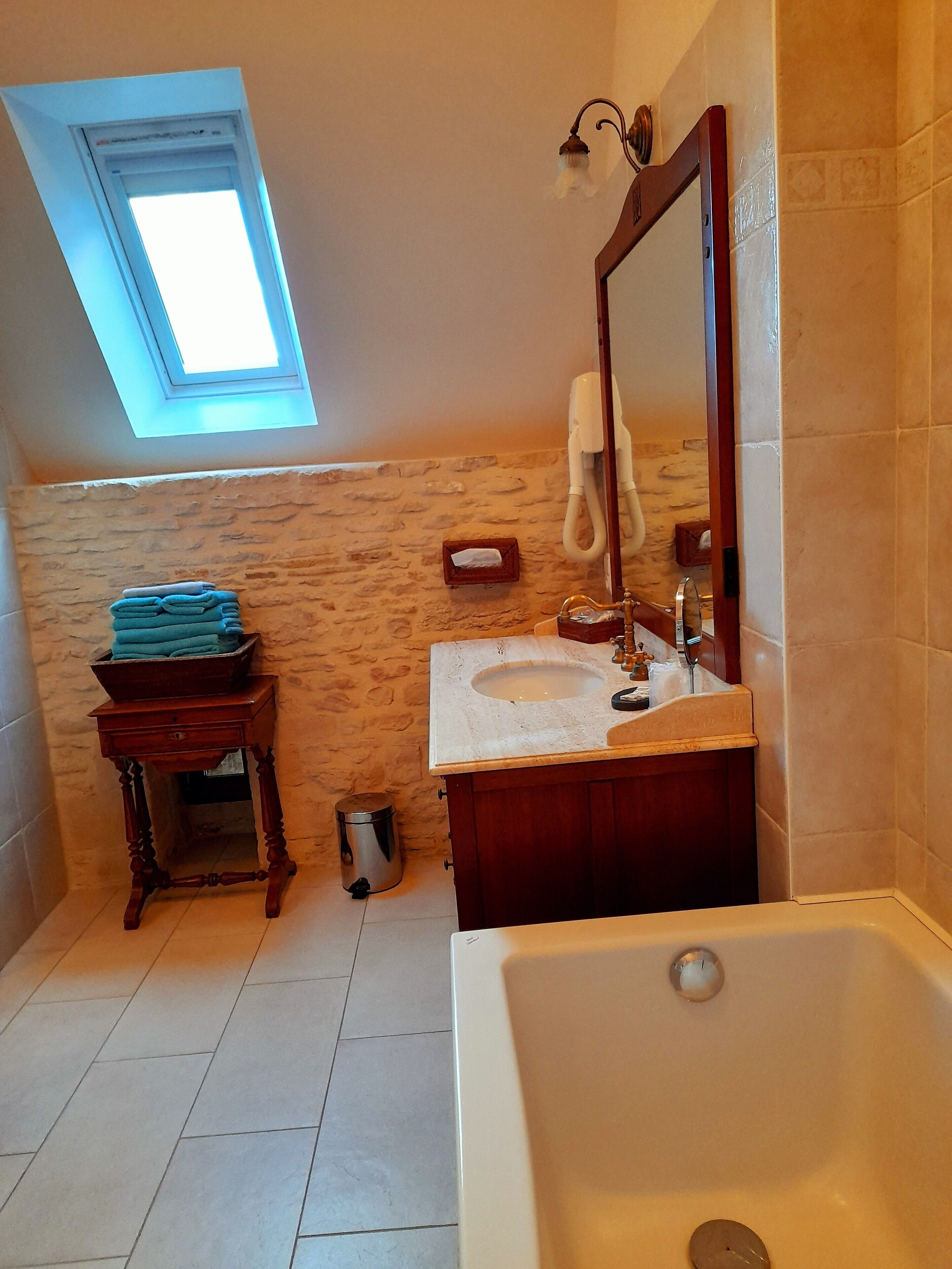 A bathroom with a skylight, a sink, a mirror, a bathtub, a wooden stool, and a trash can.