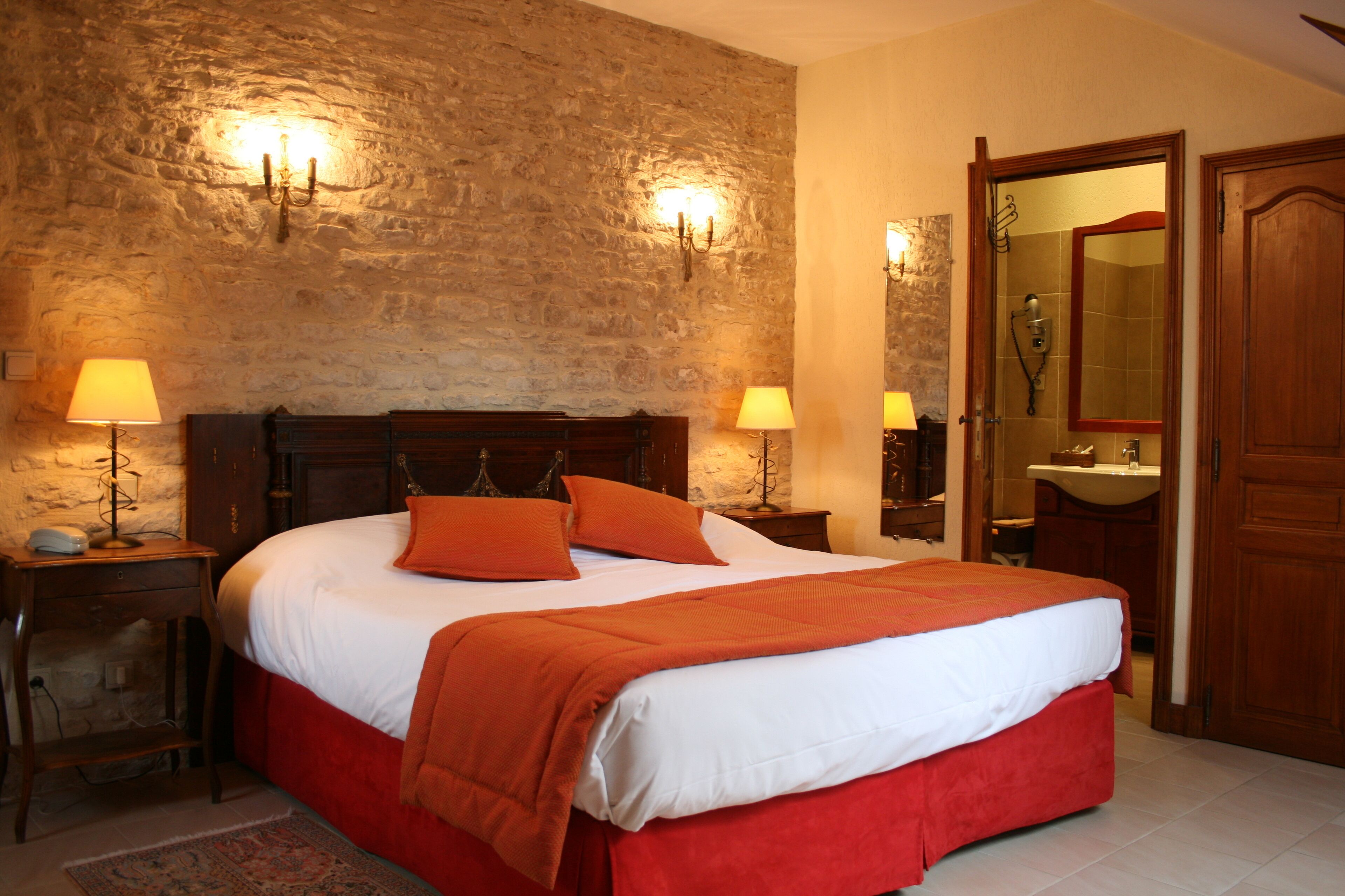 Una camera da letto con parete in pietra, un letto di legno con biancheria rossa, due lampade da comodino, un comodino, uno specchio e una porta in legno.