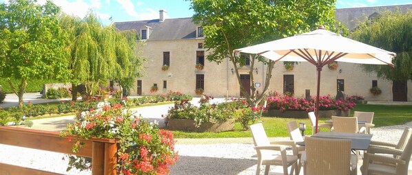 An outdoor patio area with wicker furniture and a large umbrella, surrounded by well-maintained flower beds and a stone building in the background. - Hotel Manoir de Mathan (Crepon)