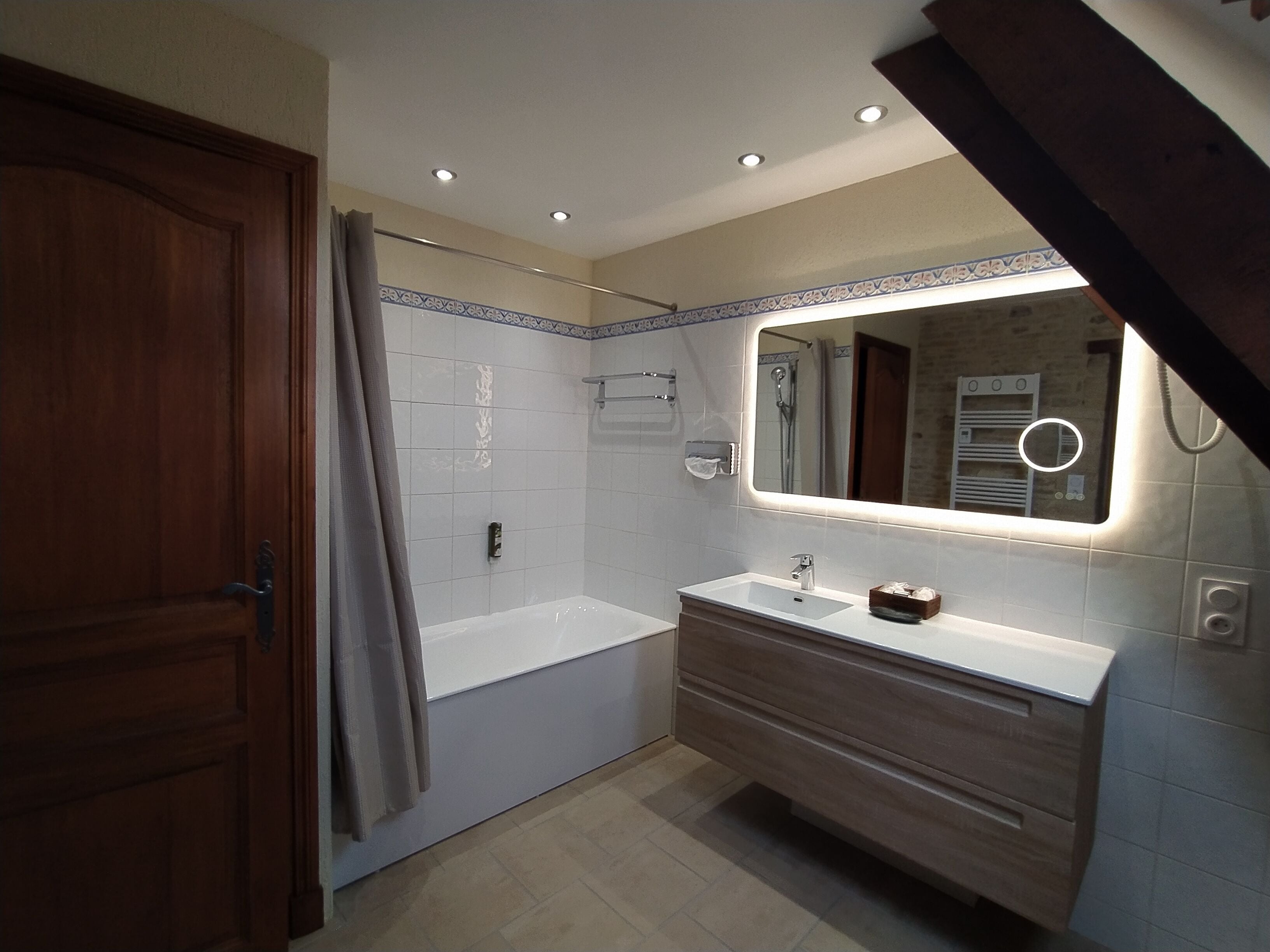 A modern bathroom with a wooden door, a shower, a vanity with a mirror, and recessed ceiling lights.