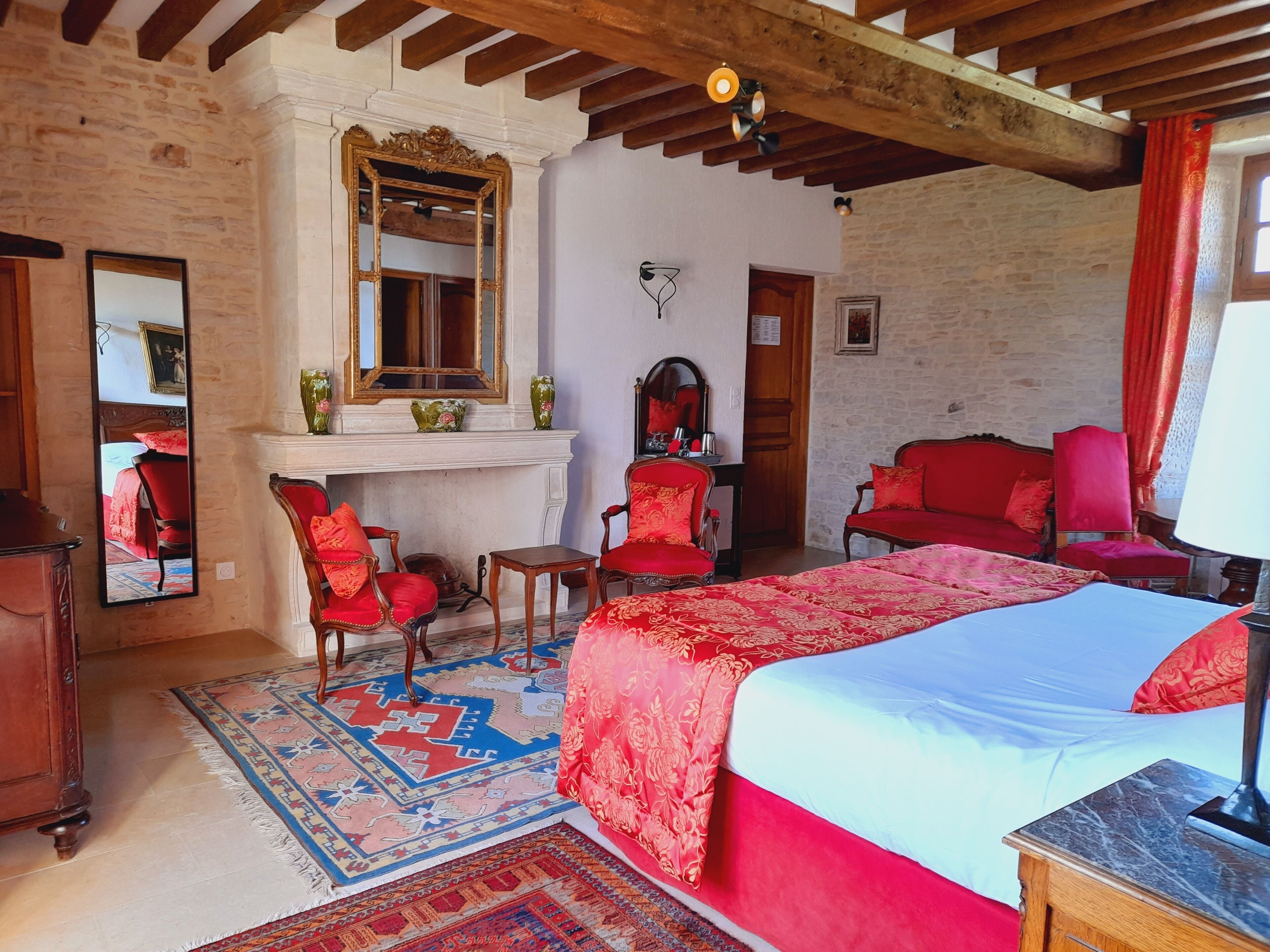Une chambre avec un grand lit, des fauteuils à revêtement rouge, une cheminée et un mur en pierre.