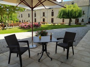 An outdoor patio with two wicker chairs and a table, surrounded by a gravel path and a garden with flowers and a tree. - Hotel Manoir de Mathan (Crepon)