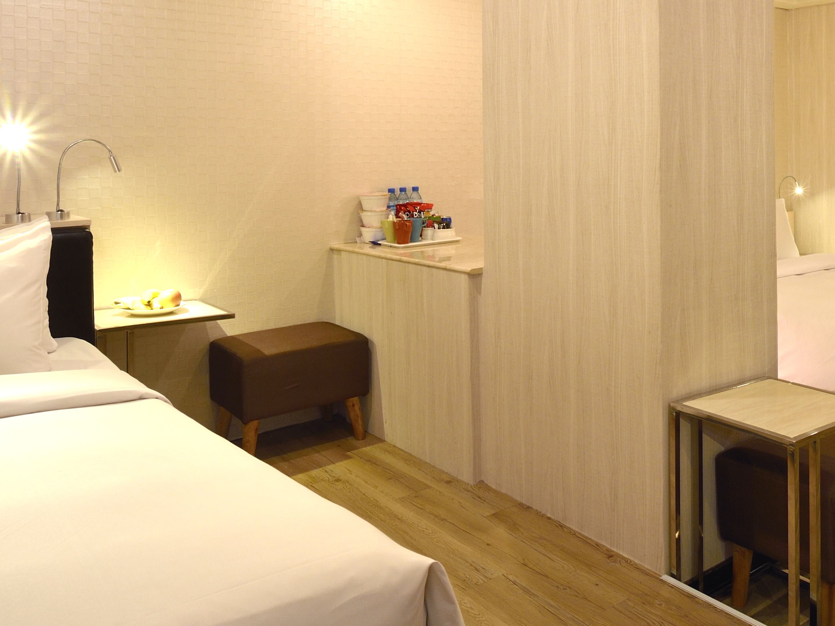 Quarto de hotel com cama, uma mesinha com abajur, um banco e prateleira com artigos de higiene pessoal.