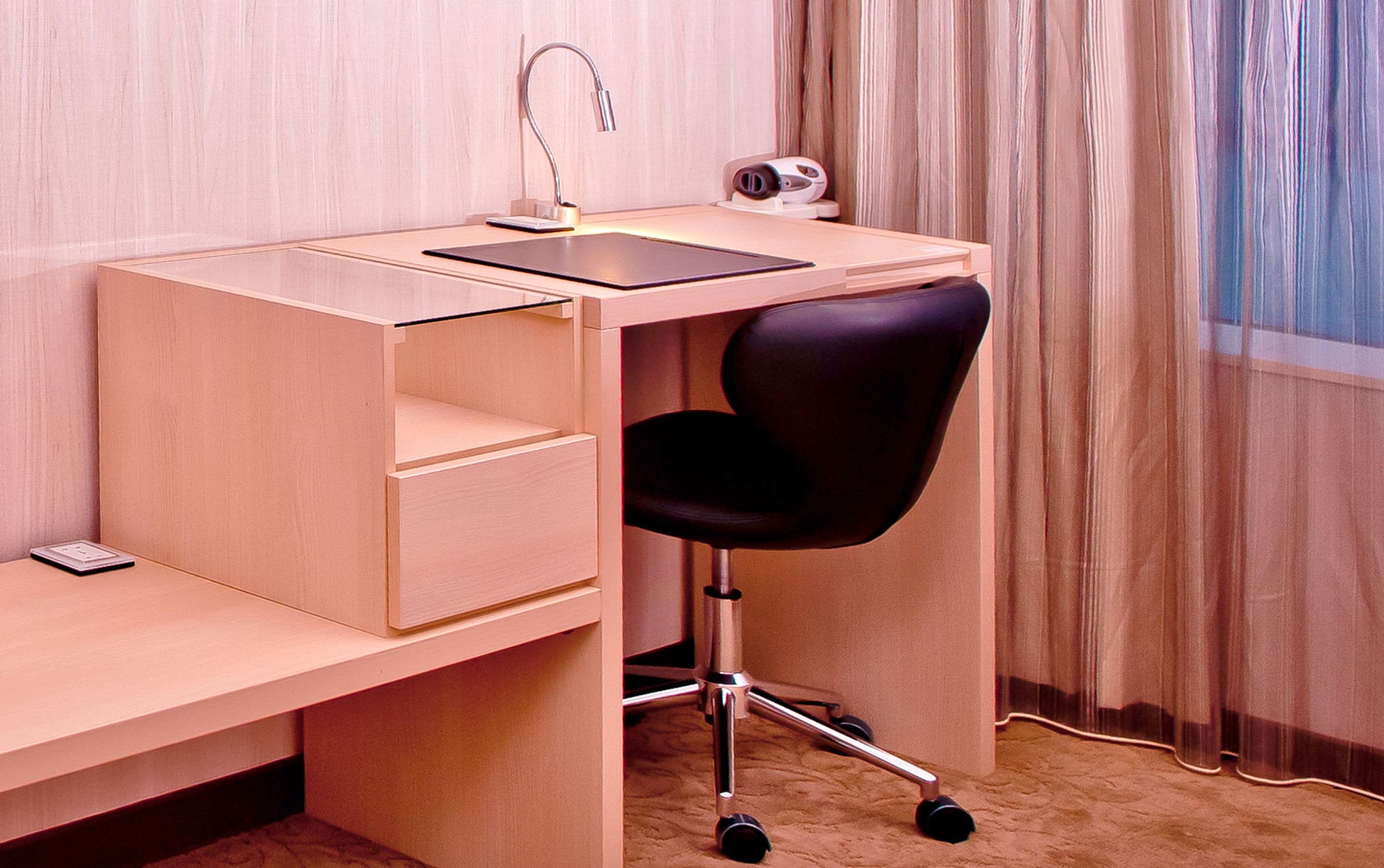 A wooden desk with a black chair, a small shelf, a desk lamp, and a security camera.