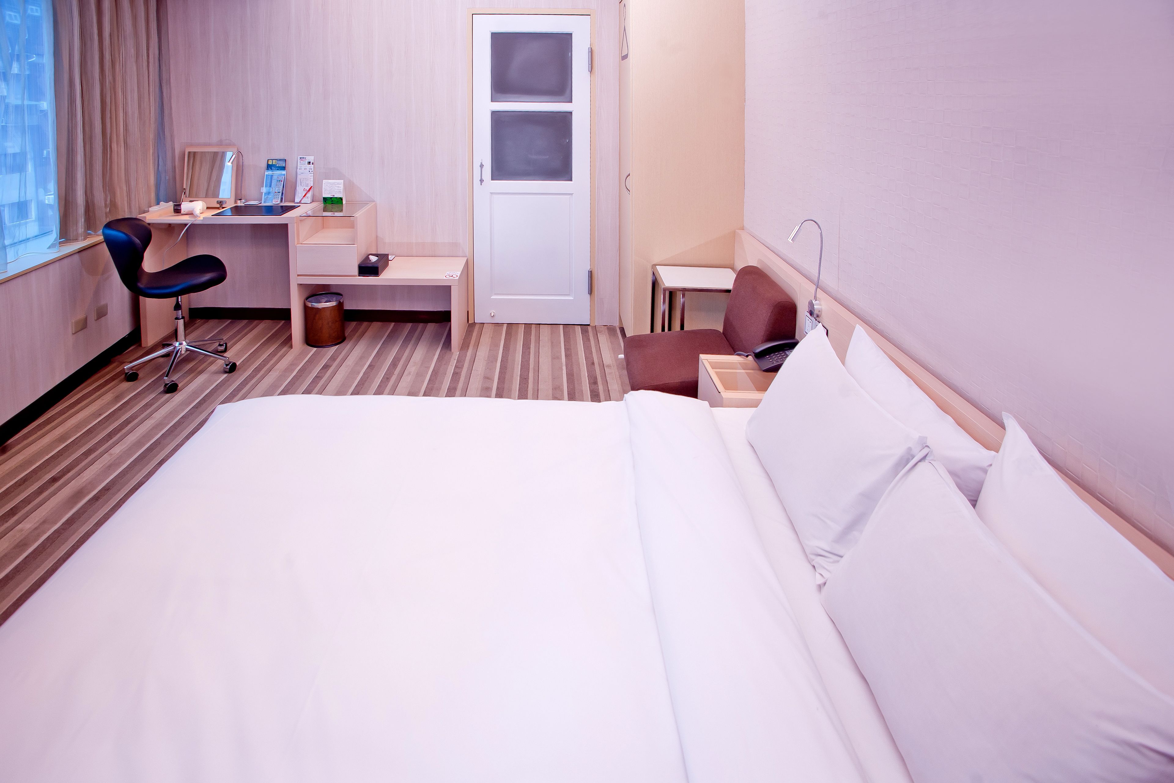 Una habitación de hotel con una cama, un escritorio con una silla, una mesita y un cubo de basura.