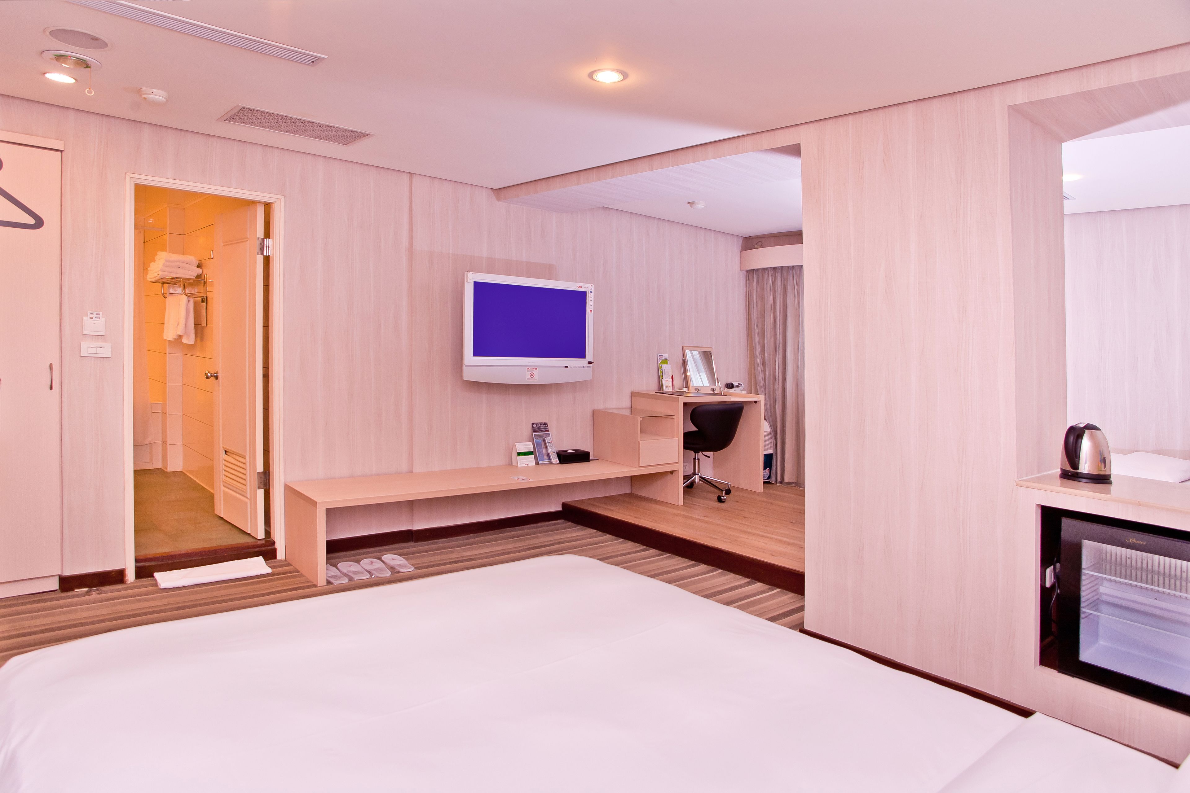 A hotel room with a bed, a desk with a chair, a television, a mirror, and a minibar.