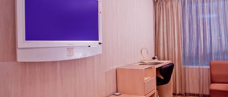 A hotel room with a flat-screen TV mounted on the wall, a desk with a chair, a bed, and a window with curtains.