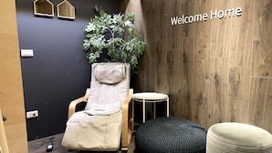 A room with a reclining chair, a small round table, a black ottoman, a potted plant, and a 'Welcome Home' sign on the wall.