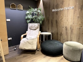 A room with a reclining chair, a small round table, a black ottoman, a potted plant, and a 'Welcome Home' sign on the wall.