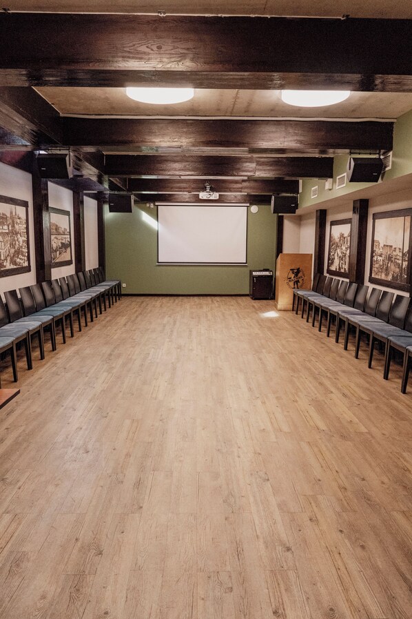 A conference room with a wooden floor, rows of chairs, a projector screen, and framed pictures on the walls.
