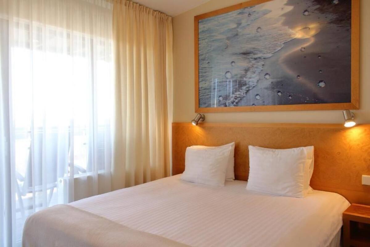 Ett hotellrum med en stor säng, vita sängkläder, en tavla på väggen och ett fönster med gardiner.