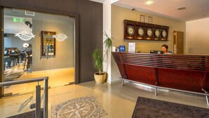 A hotel reception area with a glass door entrance, a reception desk, a clock, and a potted plant. - Hotel Kolumbs (Liepaja)