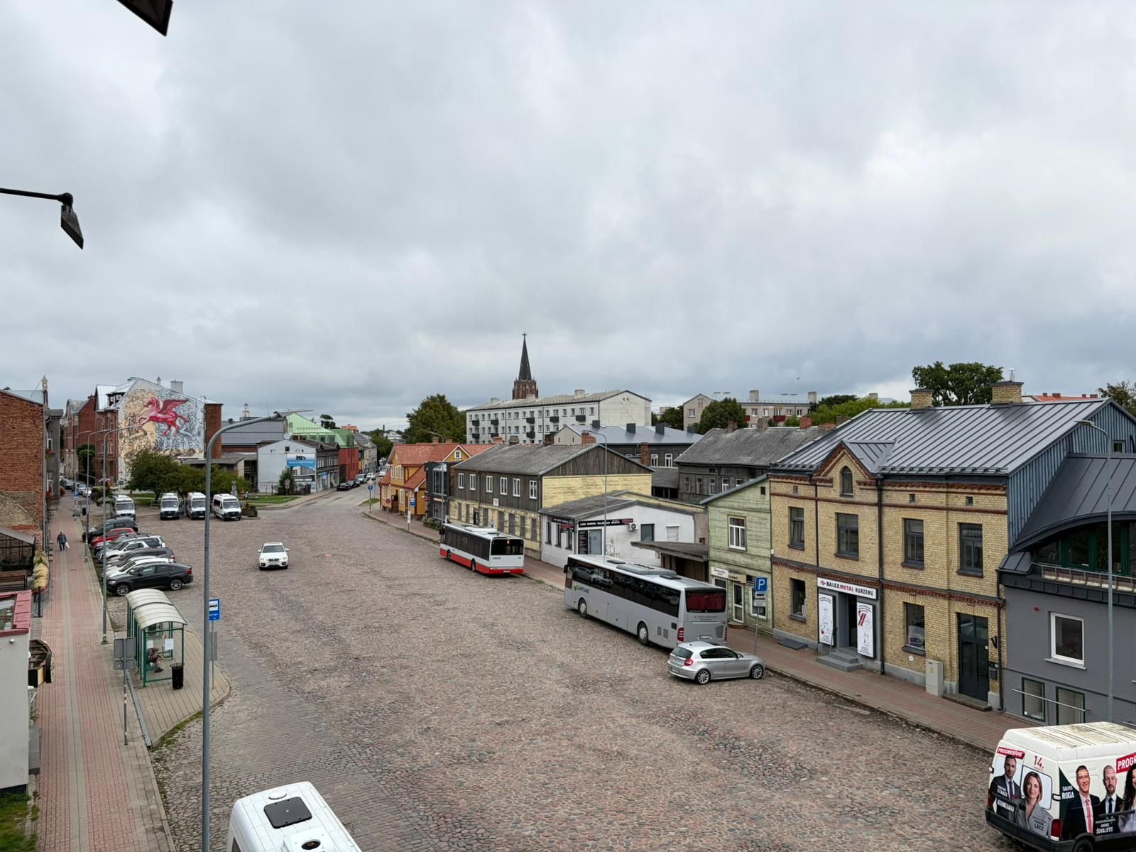 A cobblestone street lined with buildings, a bus, and parked cars.