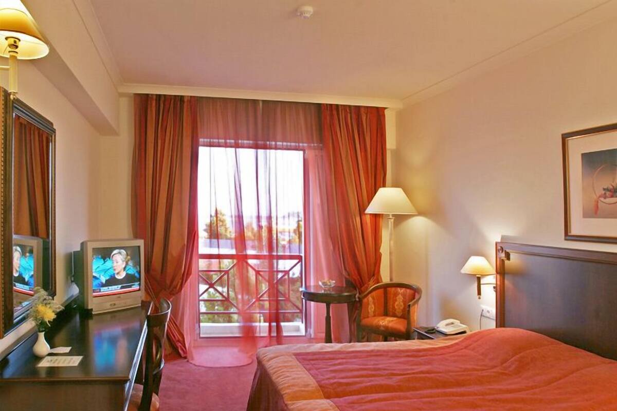 Quarto de hotel com cama, secretária com televisão, cadeira, mesa e varanda com cortinas.