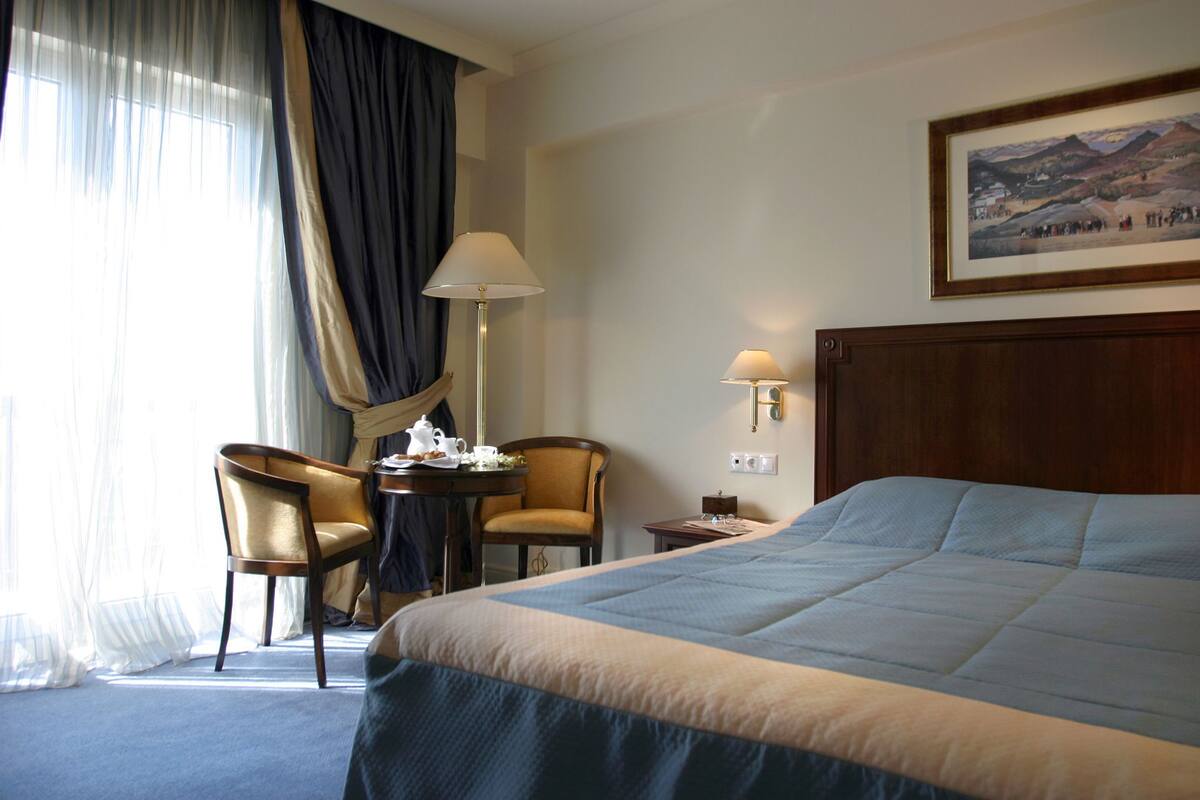 Quarto de hotel com uma cama grande, uma escrivaninha com cadeira, uma luminária e um quadro na parede.