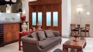A spacious living area with a sofa, a coffee table, and a dining table with chairs. There is a reception desk with a sign that reads "Coffee Shop." The room has a high ceiling with recessed lighting and a large window. - Volos Palace (Volos)