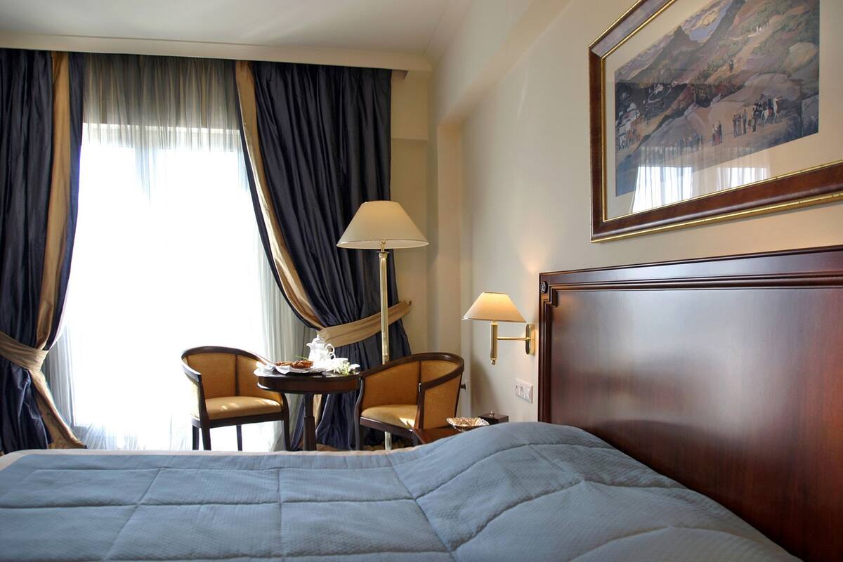 Quarto de hotel com uma cama grande, duas poltronas, uma mesa pequena com um tabuleiro de comida, uma lâmpada e um quadro na parede.