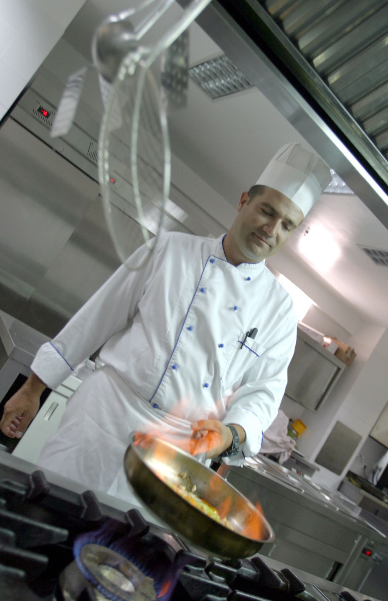 Un chef con uniforme blanco cocina en una cocina profesional equipada con electrodomésticos de acero inoxidable y una estufa a gas.