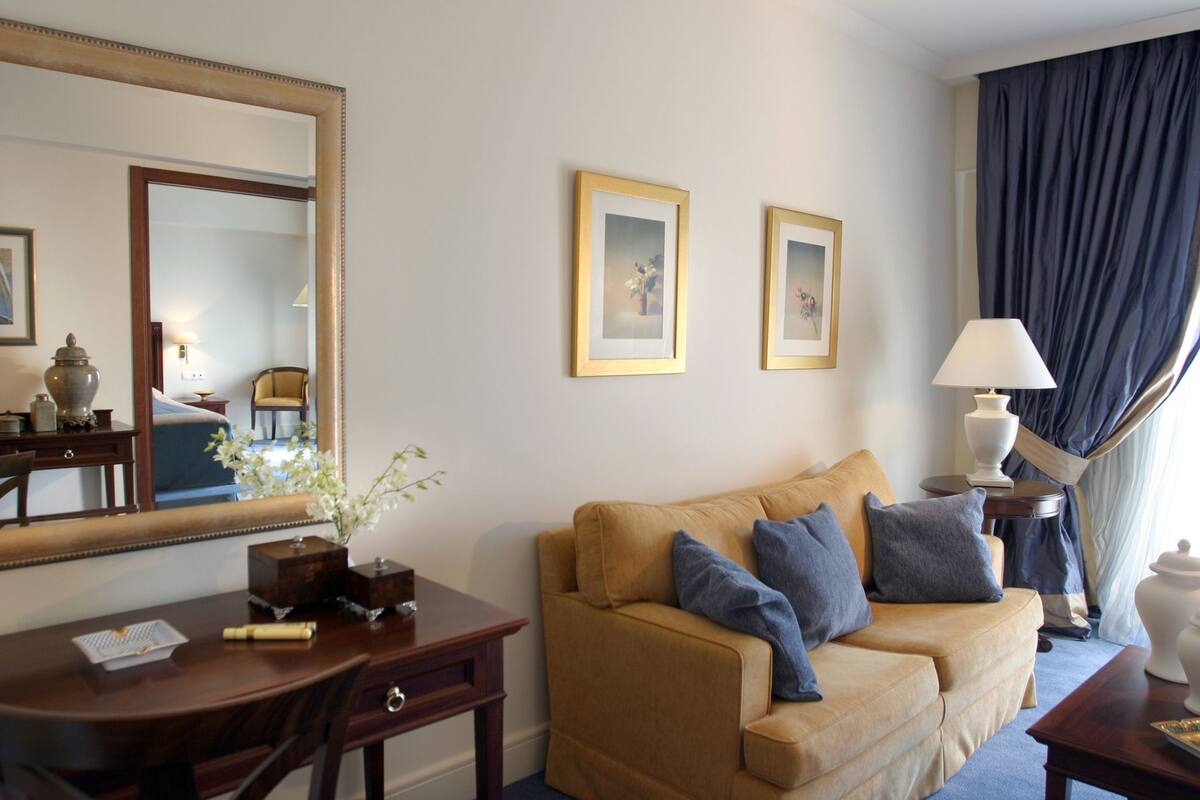 Quarto de hotel com sofá bege, escrivaninha de madeira, espelho, duas fotos emolduradas, abajur e janela com cortinas.