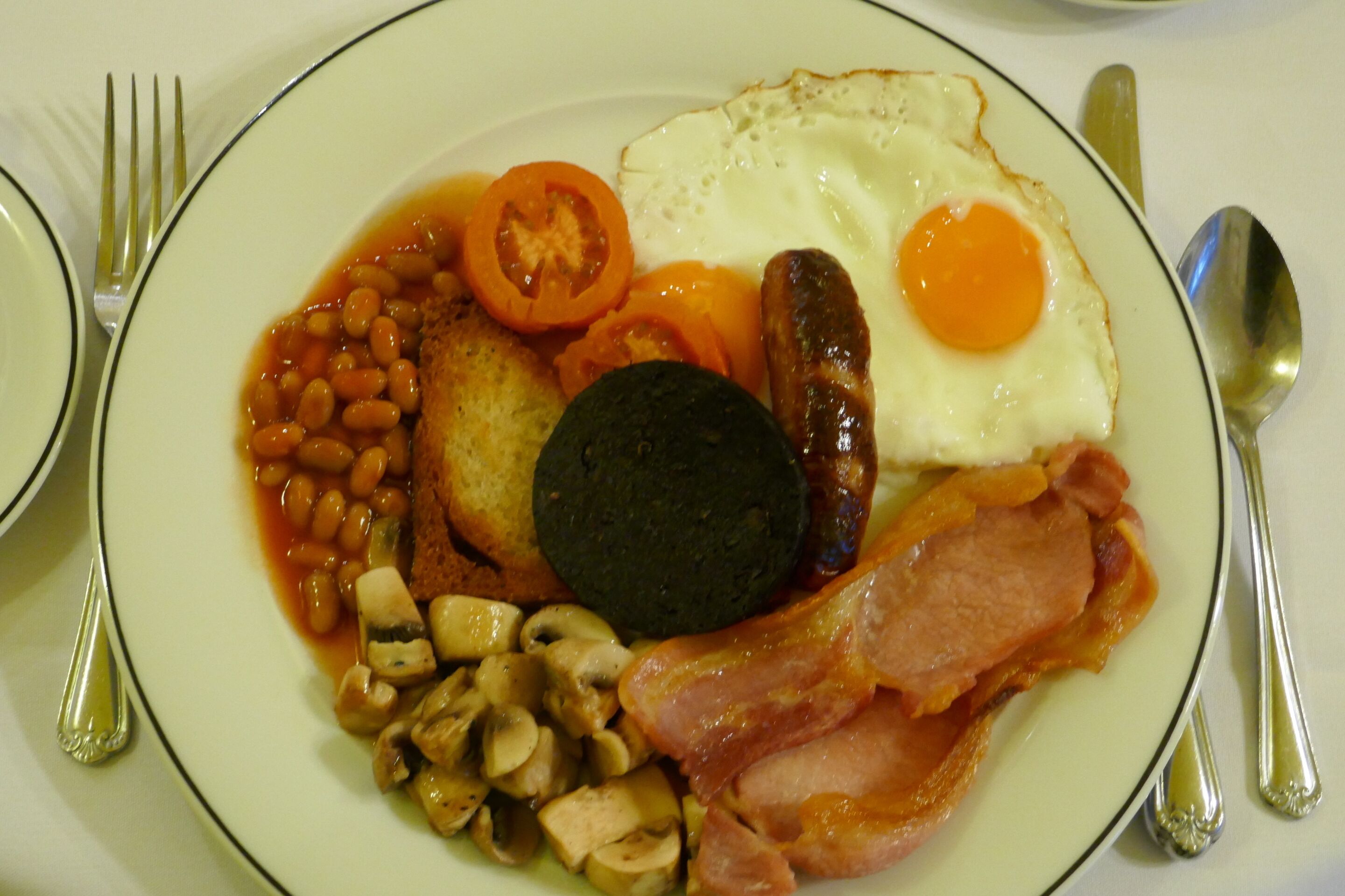 A plate of breakfast food including eggs, sausages, bacon, mushrooms, beans, tomatoes, and toast.