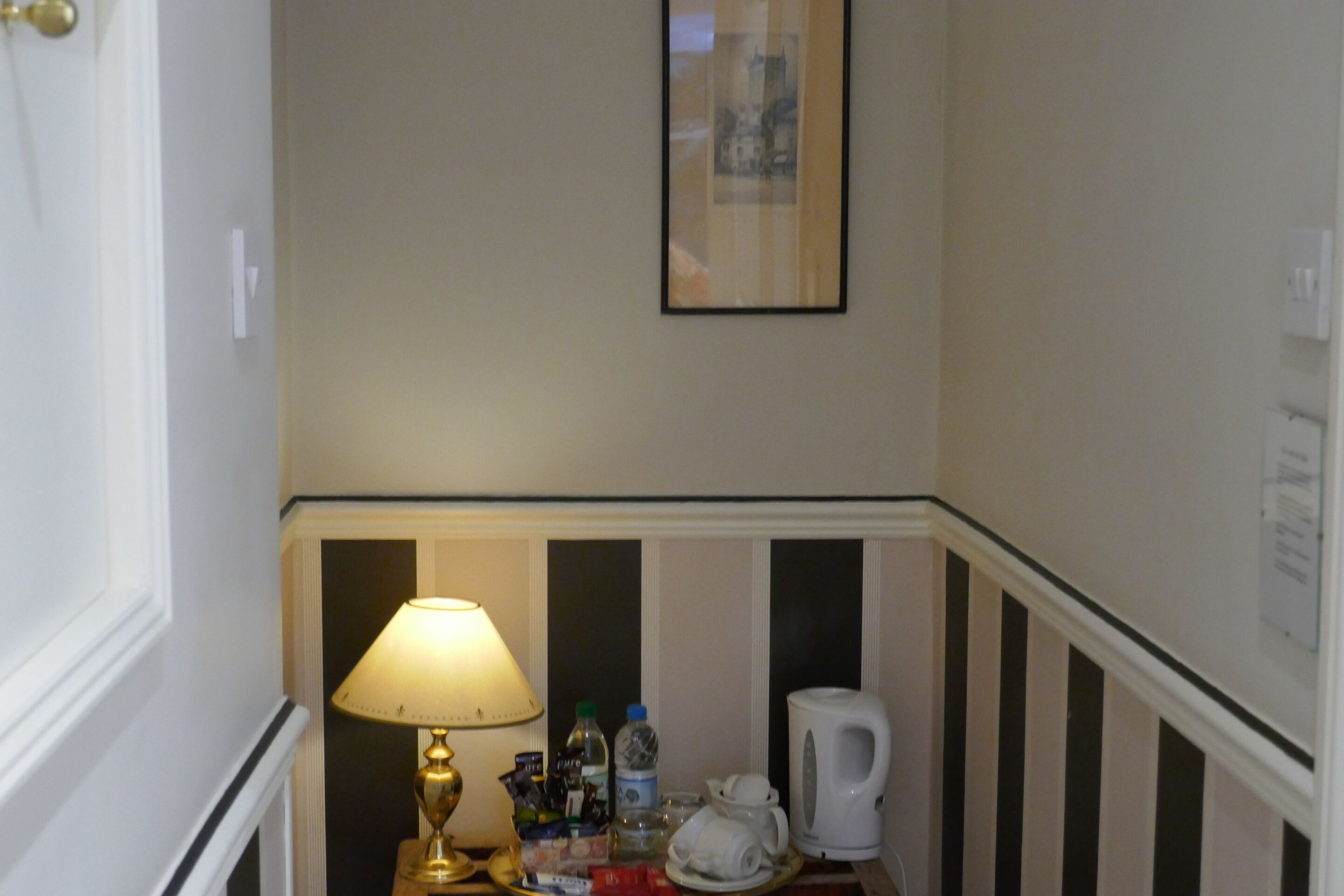 Habitación de hotel con mesita de noche, lámpara, teléfono, cafetera y un cuadro en la pared.