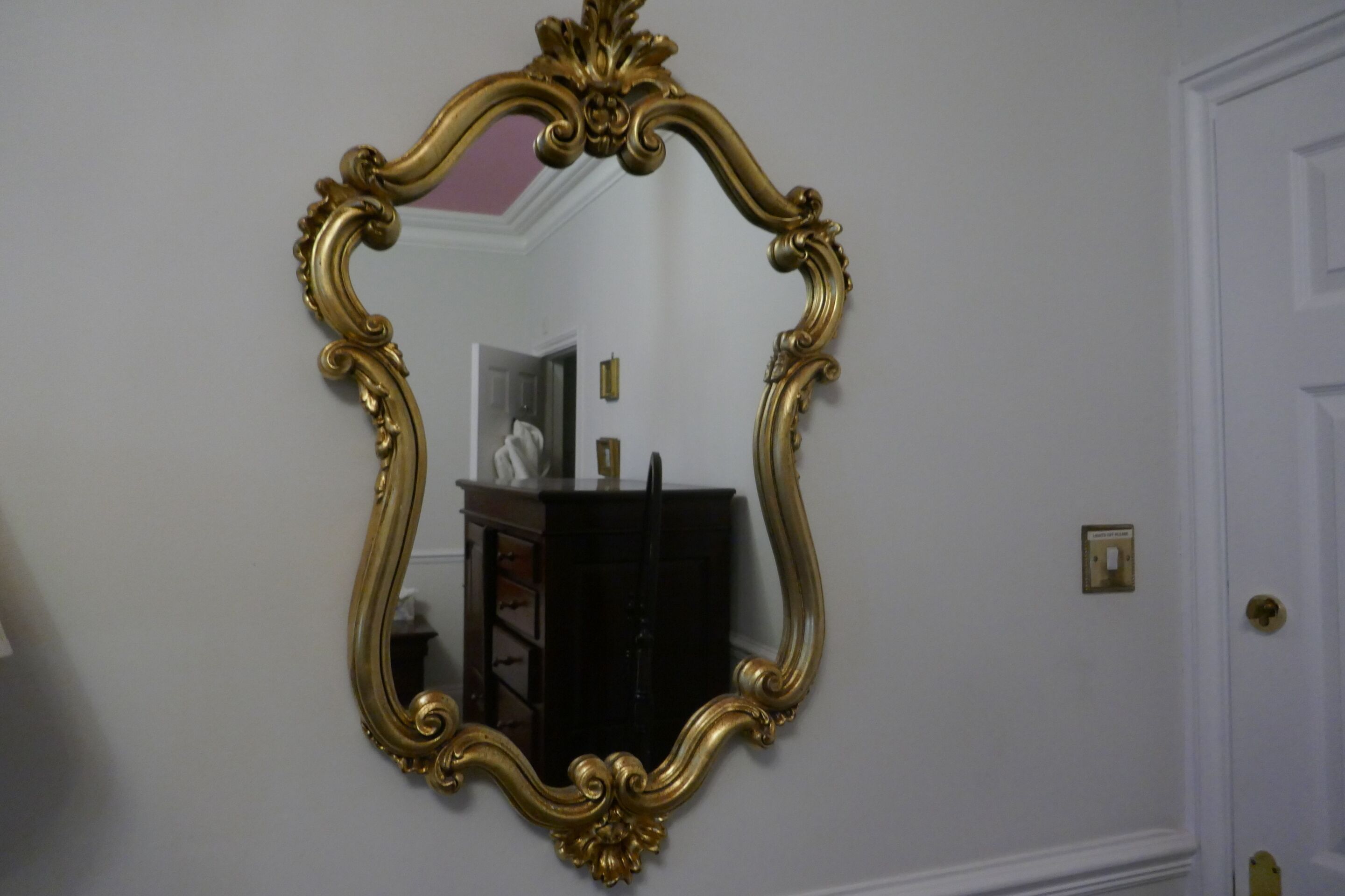 A room with a large ornate mirror on the wall, a door to the right, and a small table with a lamp in the background.