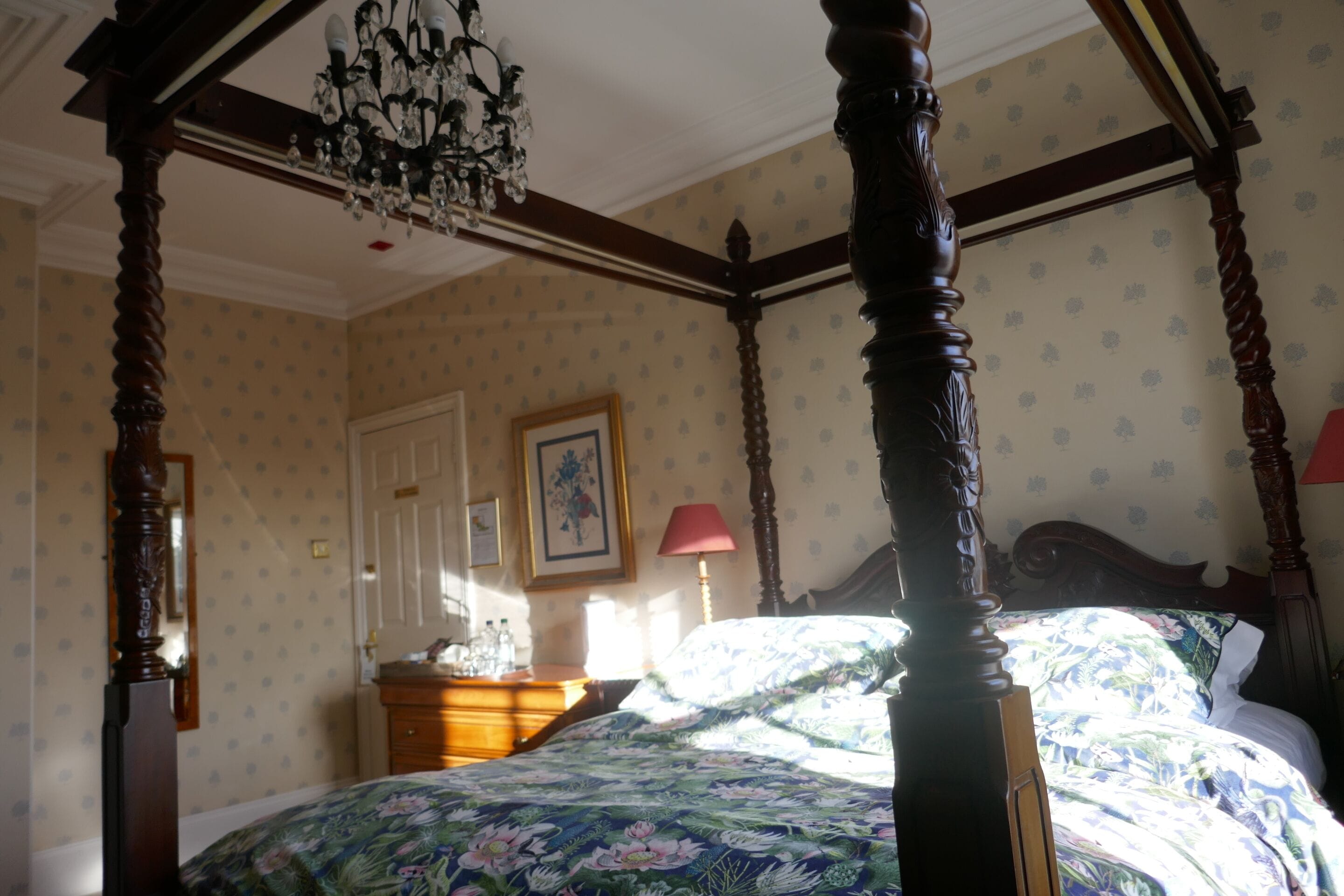 A four-poster bed with a floral bedspread, a wooden dresser, a mirror, and a chandelier.