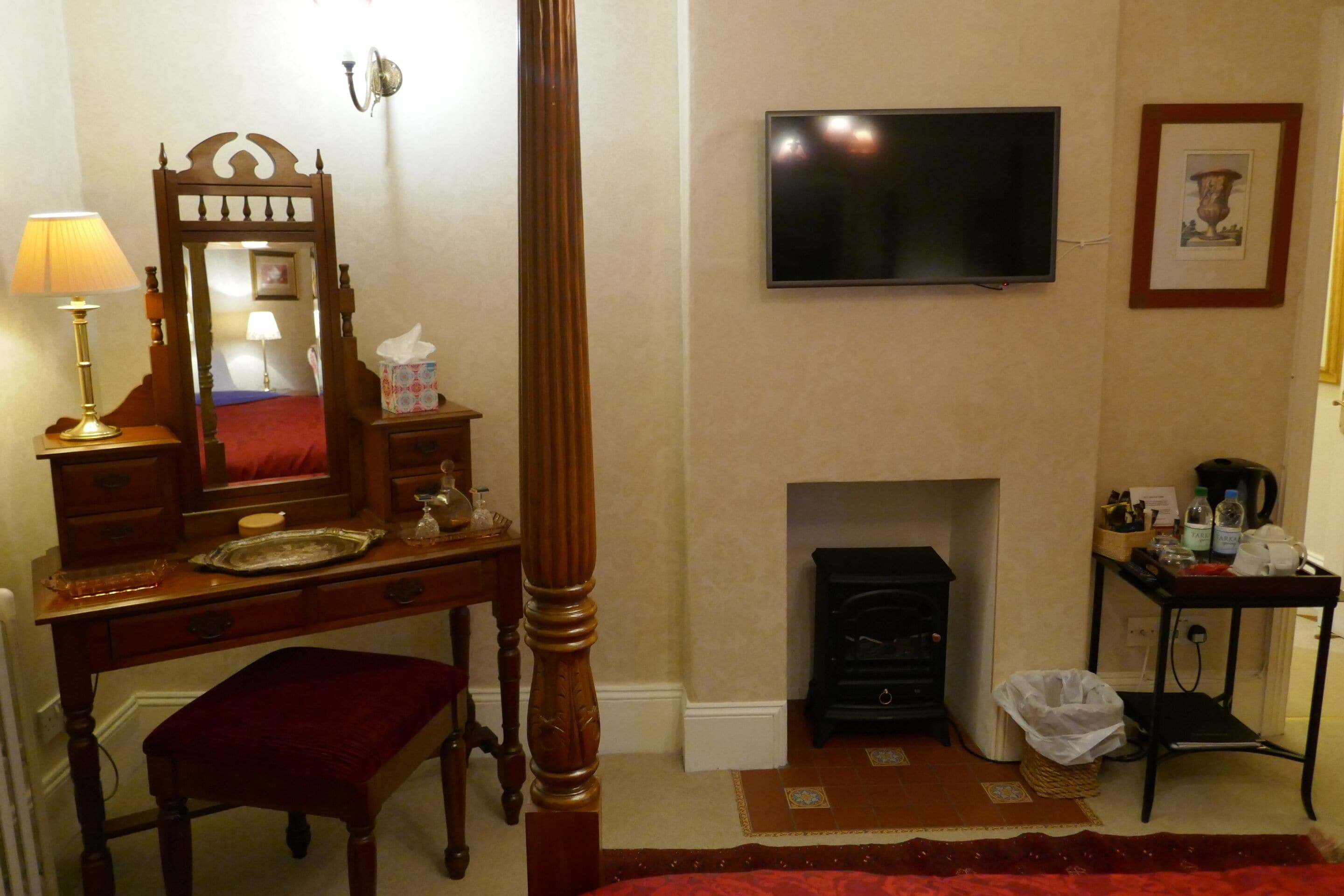 Un dormitorio con una cama de madera, una mesita de noche con lámpara, un espejo, un televisor en la pared y una chimenea.