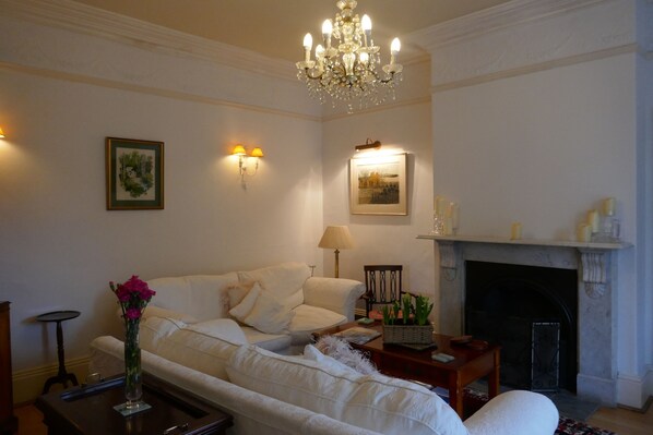 A living room with a white sofa, a chandelier, a fireplace, and wall-mounted lamps. - Kerrington House (Axminster)