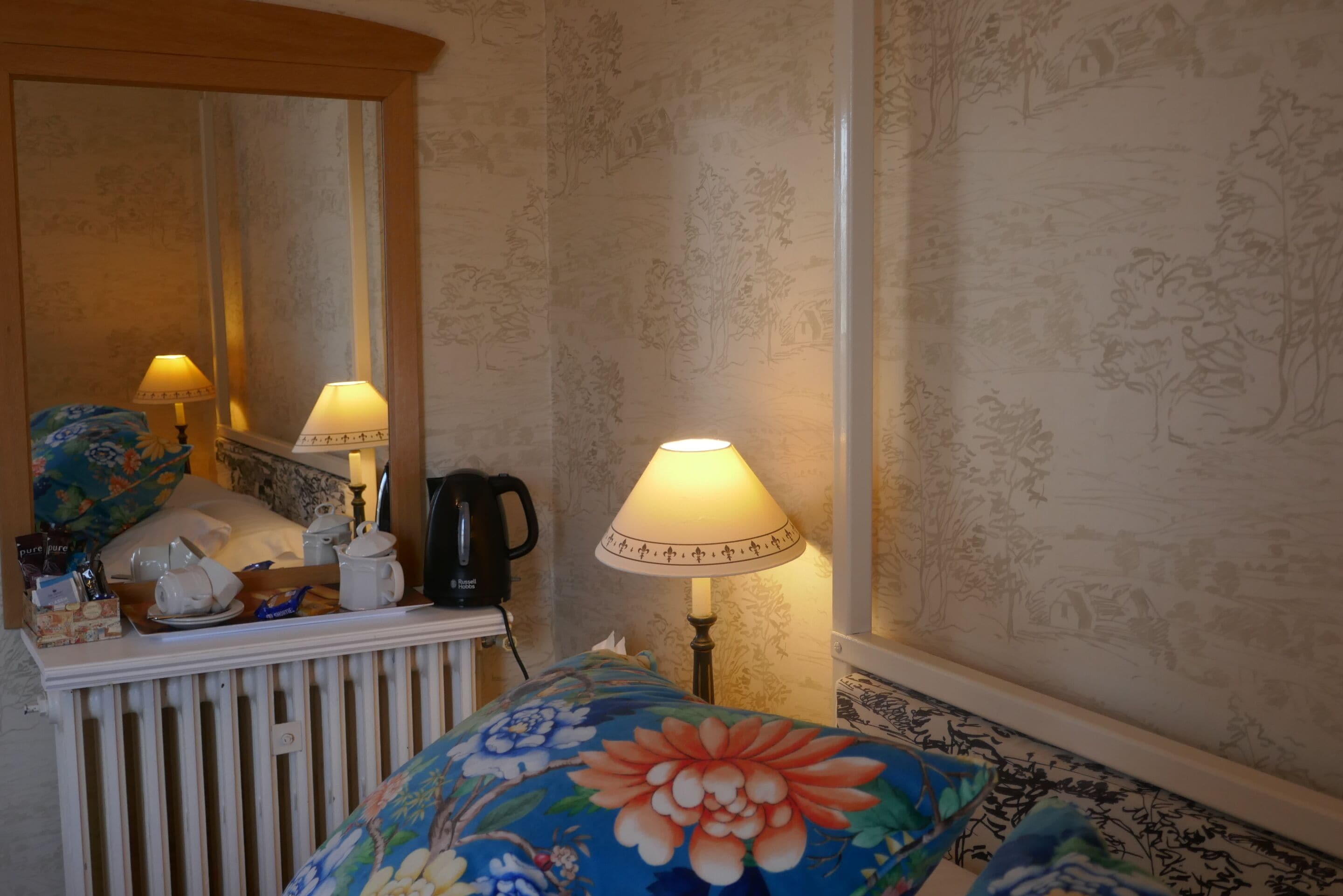 Ein Hotelzimmer mit Bett, Nachttisch mit Lampe, Wasserkocher und Spiegel.