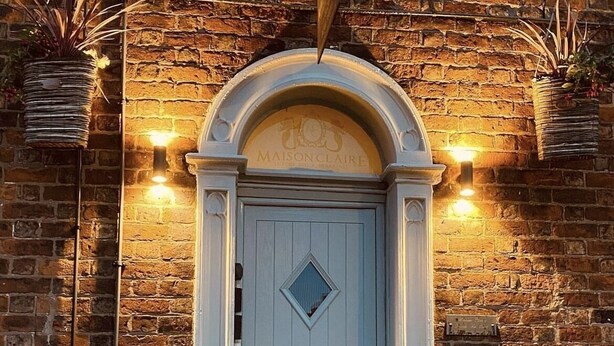 A light blue door with a diamond-shaped window, flanked by two hanging planters, set against a brick wall with a stone arch above. - Maison Claire (Chester)