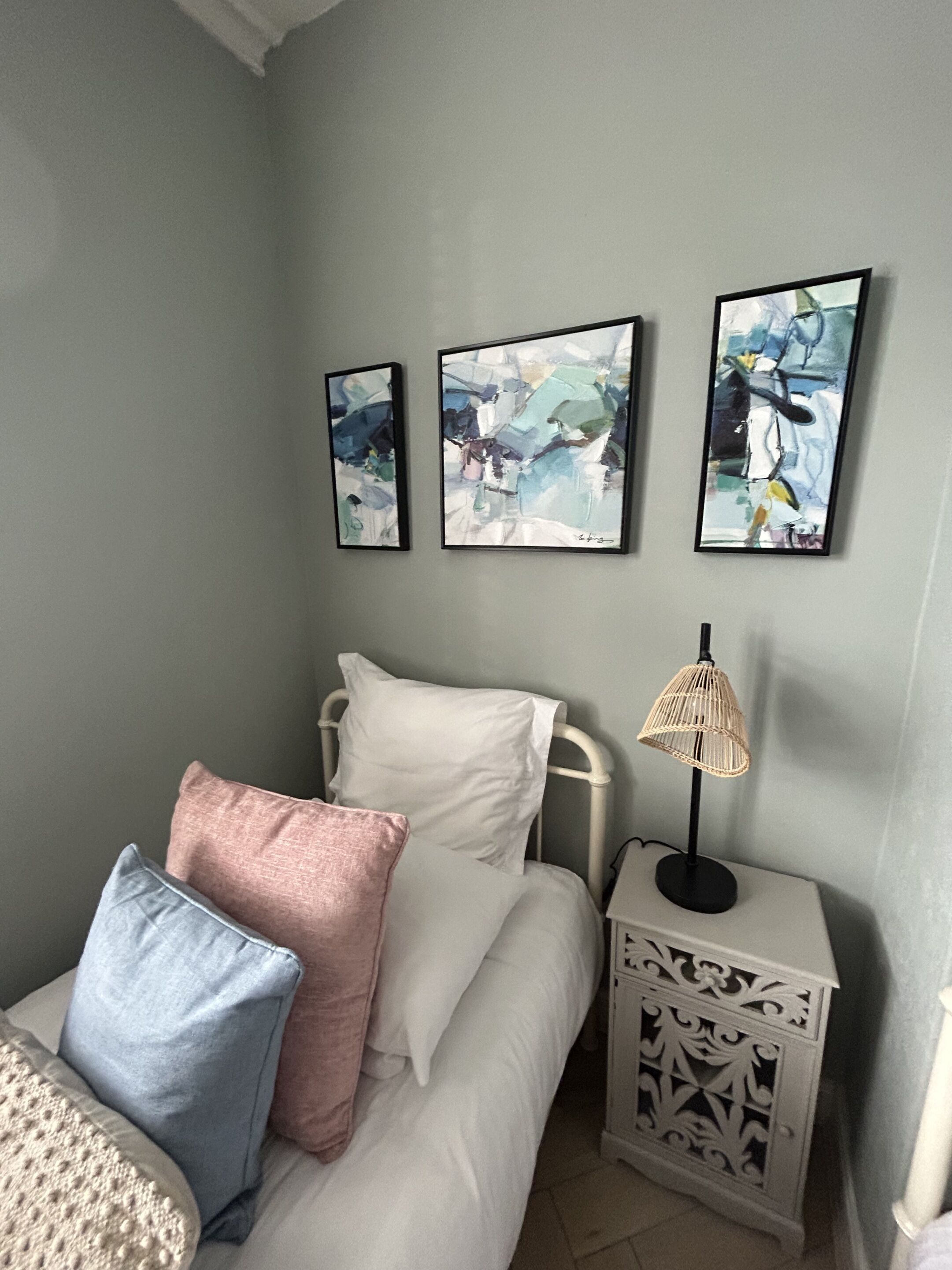 A bedroom with a bed, pillows, a bedside table with a lamp, and three abstract paintings on the wall.