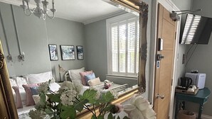 A bedroom with a large mirror, a wooden dresser, a vase with flowers, a bed with pillows, a television, and a window with blinds. - Maison Claire (Chester)