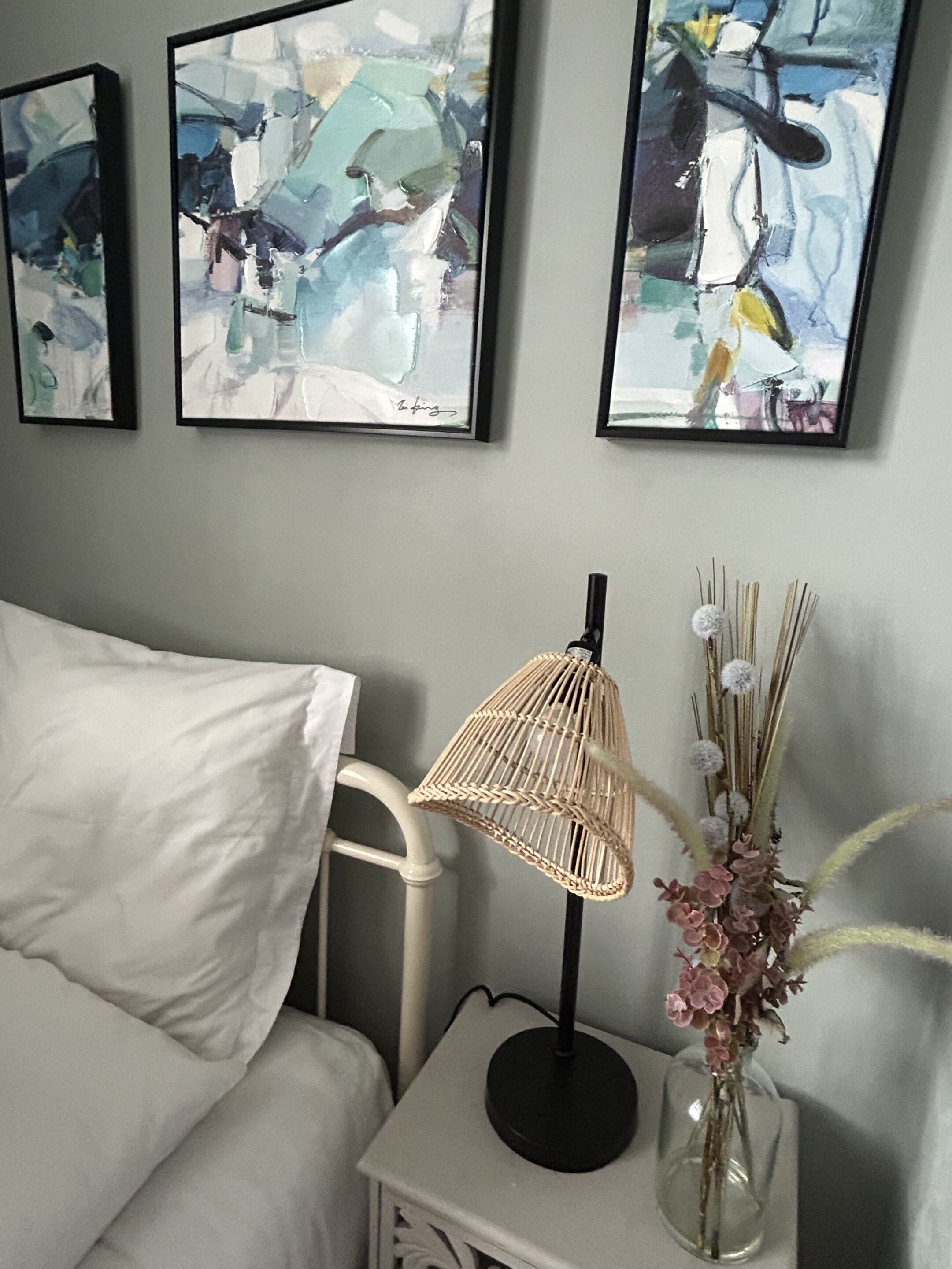 Un dormitorio con una cama blanca, una mesita de noche con una lámpara y un jarrón de flores, y pinturas abstractas en la pared.