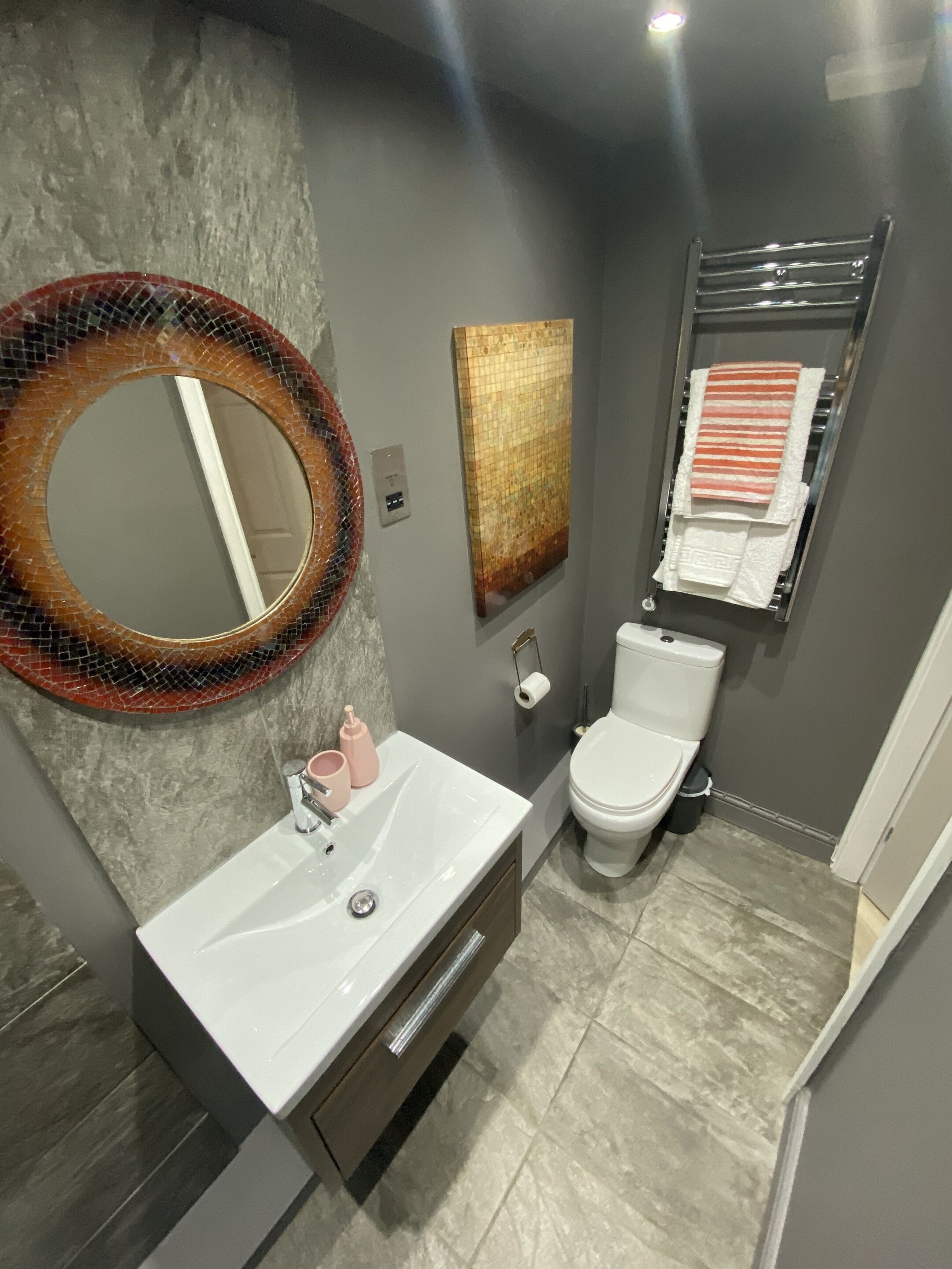 A modern bathroom with a round mirror, a white sink, a toilet, and a towel rack.