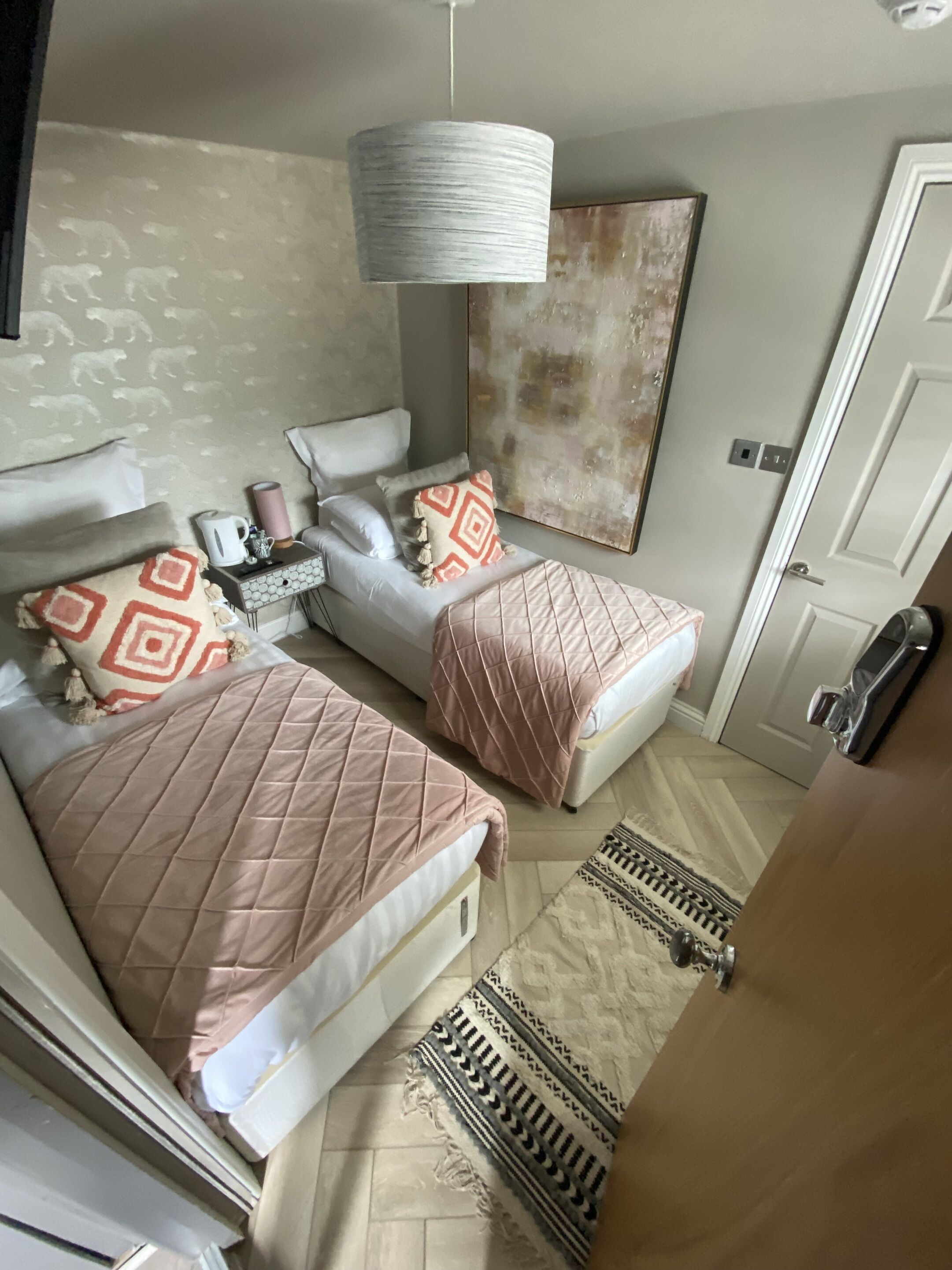 Un dormitorio con dos camas, una mesita de noche con lámpara, un cuadro en la pared y una puerta.