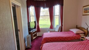 A bedroom with two beds, a desk, a chair, and a radiator. There is a window with curtains, a chandelier, and a painting on the wall. - Mackenzie Guest House (Edinburgh)