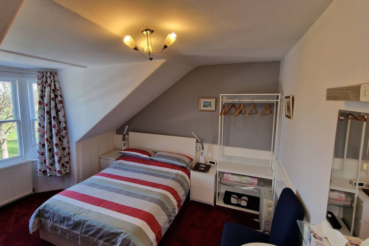 Quarto com teto inclinado, cama com colcha listrada, mesa de cabeceira com abajur, espelho e guarda-roupa com cabides.