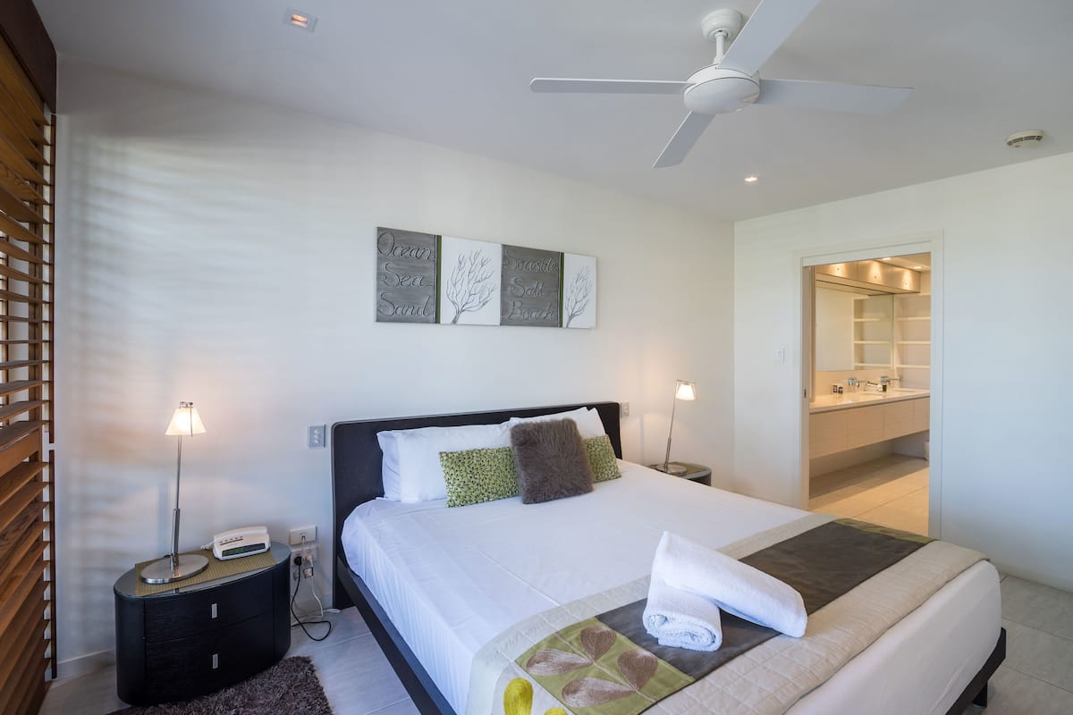 A modern hotel room with a bed, bedside tables, a ceiling fan, and a bathroom visible through an open door.