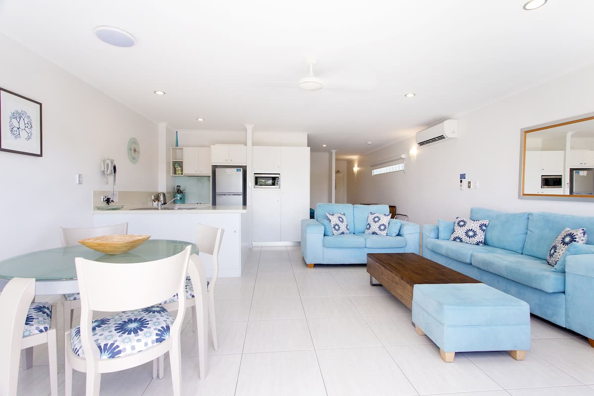 A modern living room with a blue sofa, a wooden coffee table, and a glass dining table with chairs.