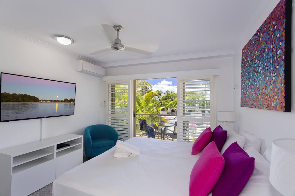 A modern hotel room with a bed, a TV, a ceiling fan, and a balcony with a view of the outdoors.