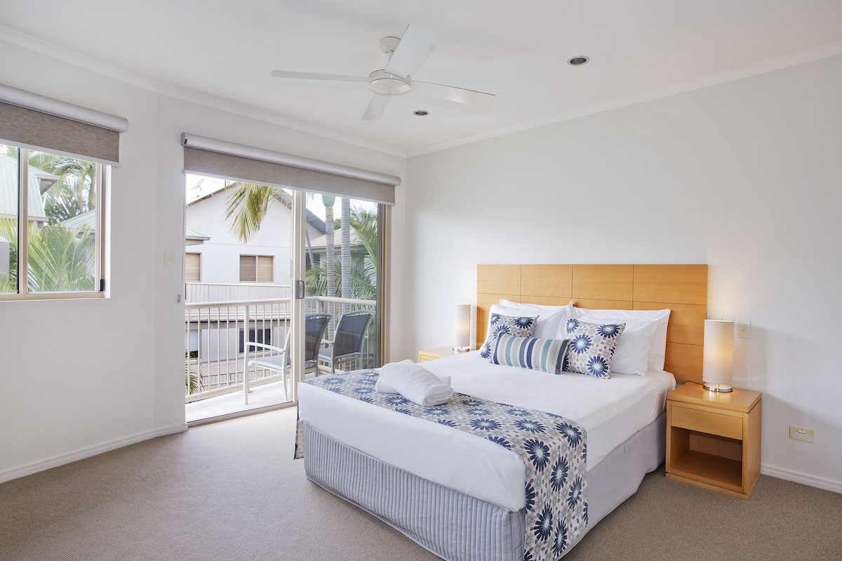 A hotel room with a large bed, a wooden headboard, a nightstand with a lamp, and a balcony with chairs.