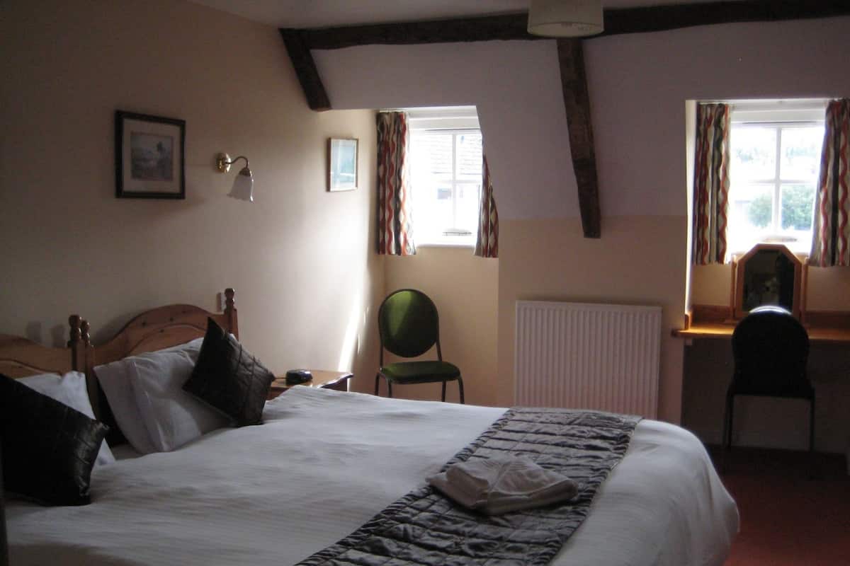 A bedroom with a large bed, a desk, and a chair. There are windows with curtains, a radiator, and framed pictures on the wall.