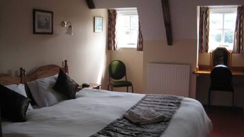 A bedroom with a large bed, a desk, and a chair. There are windows with curtains, a radiator, and framed pictures on the wall.
