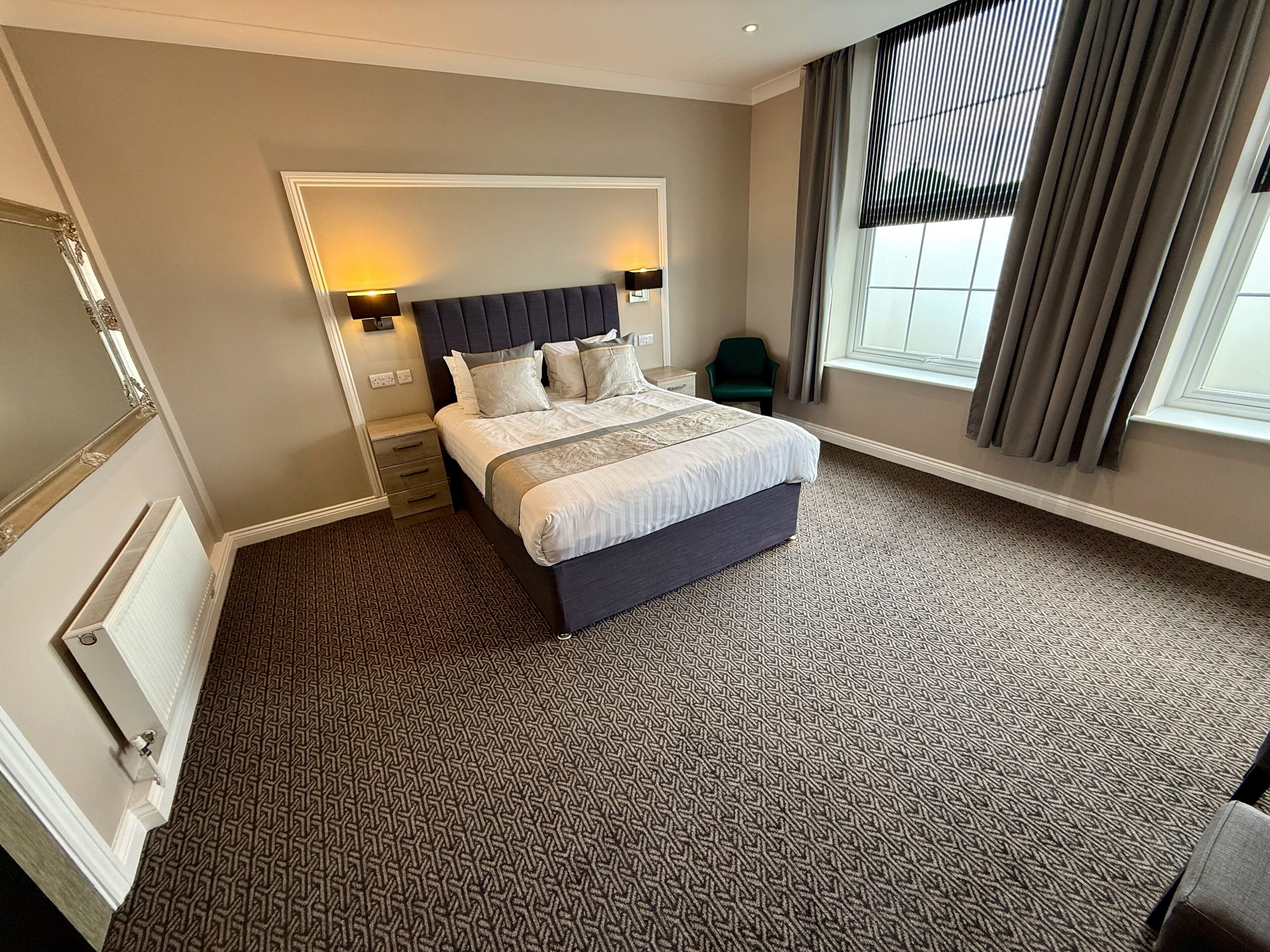 Habitación de hotel con una cama grande, una mesita de noche, una silla, una ventana con cortinas y un radiador.